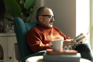 Uomo seduto in poltrona mentre legge un libro in casa, esempio di attività culturale associata al benessere mentale e all’invecchiamento attivo.