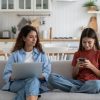 Madre e figlia adolescenti sedute sul divano mentre usano laptop e smartphone in un contesto di educazione digitale familiare.