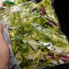 Mano che tiene una confezione di insalata in busta pronta al consumo nel reparto refrigerato di un supermercato.