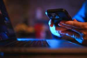 Persona che utilizza uno smartphone accanto a un computer portatile in ambiente serale. App di incontri e piattaforme digitali possono facilitare l’organizzazione di incontri sessuali e l’accesso a contesti associati al chemsex.