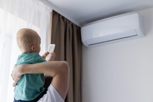 Adulto con un bambino in braccio mentre utilizza il telecomando di un condizionatore domestico. L’aria condizionata può essere usata anche in presenza di neonati e bambini, evitando sbalzi termici e getti d’aria diretti.