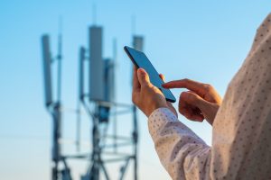 Persona che utilizza uno smartphone vicino a un’antenna 5G, immagine che rappresenta l’esposizione ai campi elettromagnetici delle reti mobili e il dibattito sui possibili effetti sulla salute.