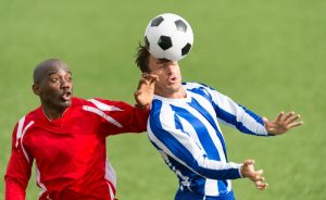 Due calciatori adulti si contendono un pallone alto con un colpo di testa durante una partita, situazione in cui possono verificarsi anche contatti tra giocatori.