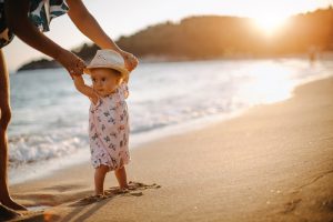 Bambina con cappello cammina sulla spiaggia al tramonto accompagnata da un adulto, esempio di esposizione al sole in orari meno caldi.