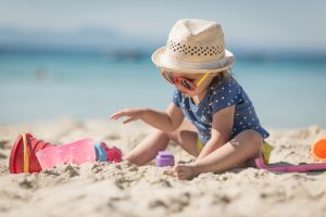Bambina con cappello, occhiali da sole e giochi sulla spiaggia, protetta dal sole con indumenti e accessori durante una giornata al mare.
