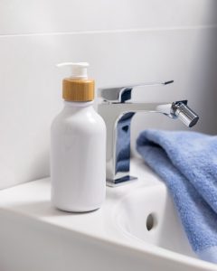 Flacone di detergente intimo accanto a un lavabo e a un bidet, immagine che richiama l'igiene quotidiana dei genitali esterni.