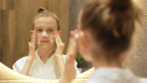Ragazza davanti allo specchio mentre applica una crema sul viso, esempio di routine cosmetica in età preadolescenziale.