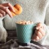 Persona con maglione che tiene in mano un biscotto e una tazza di bevanda con panna montata.